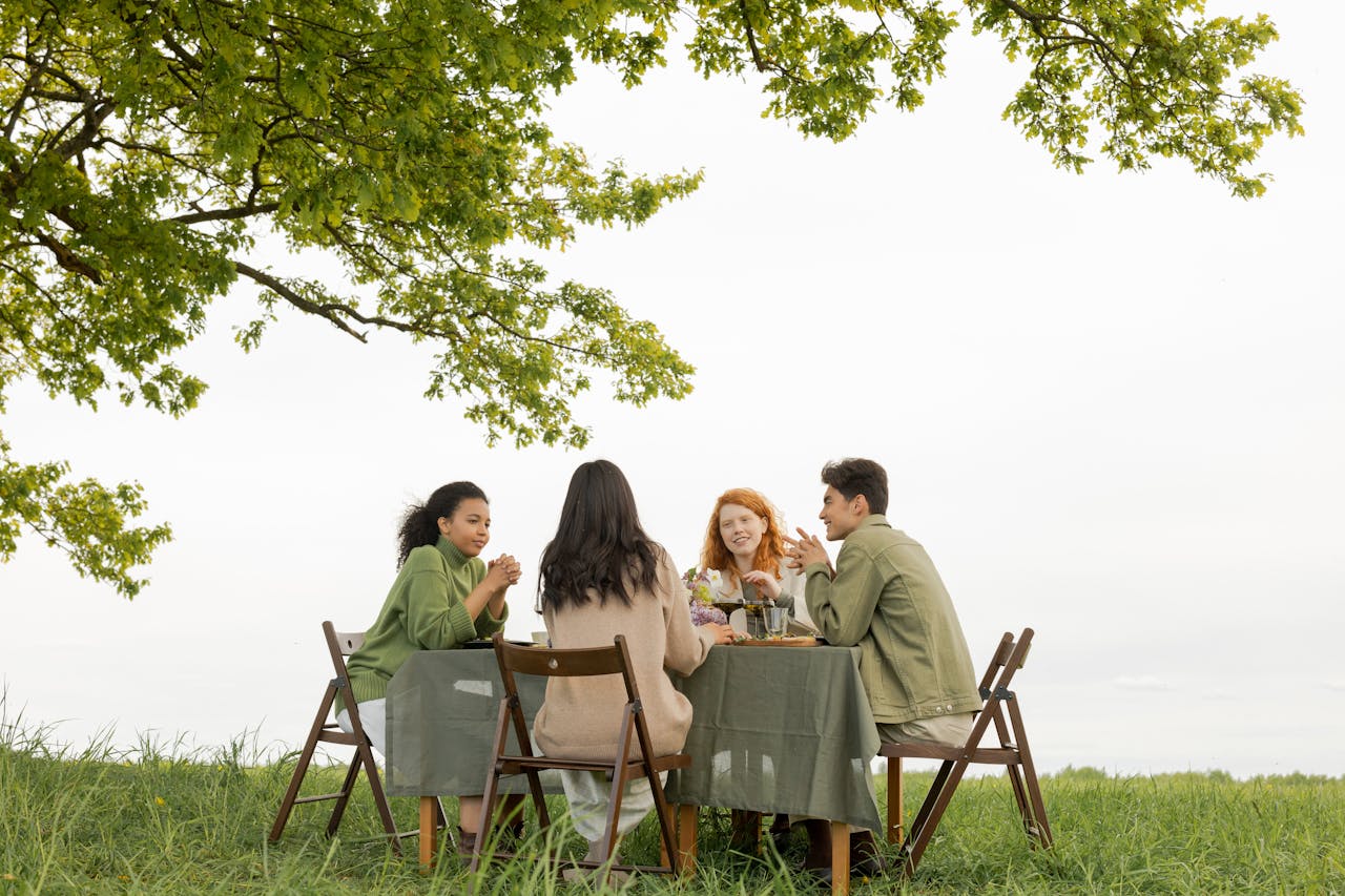 A diverse group of friends at an outdoor picnic, enjoying food and conversation under a tree.